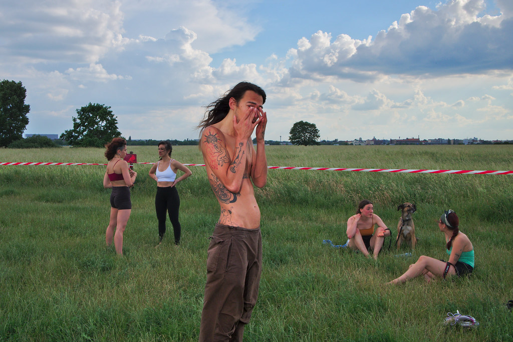 A shirtless, tattooed man wipes his face in an open field, with two women and a dog behind a line of striped barrier tape.